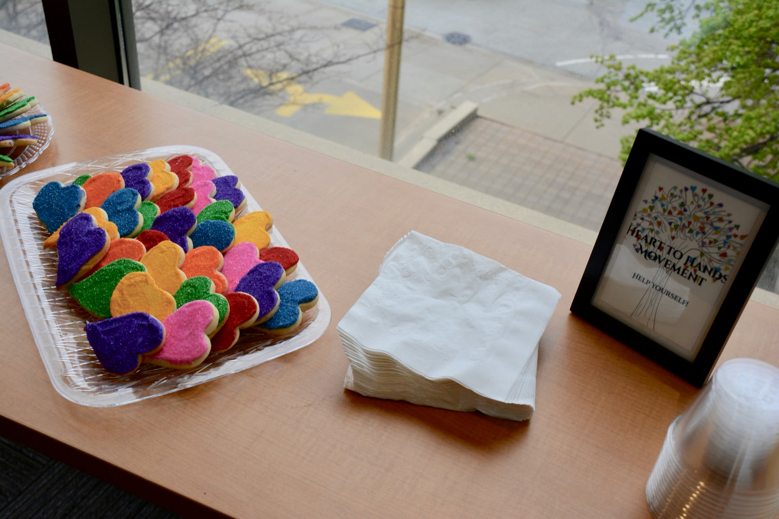 Rainbow heart-shaped cookies beside a framed Heart to Hands Movement 'Help Yourself!' sign at the book launch